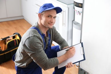 Photo of Male technician with clipboard examining broken refrigerator in kitchen