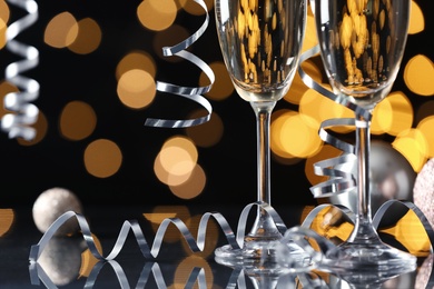 Photo of Glasses of champagne, Christmas decor and serpentine streamers against black background with blurred lights, closeup. Space for text