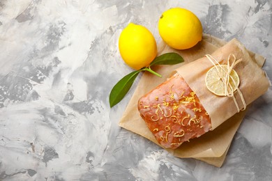 Photo of Wrapped tasty lemon cake with glaze and citrus fruits on light grey textured table, top view. Space for text