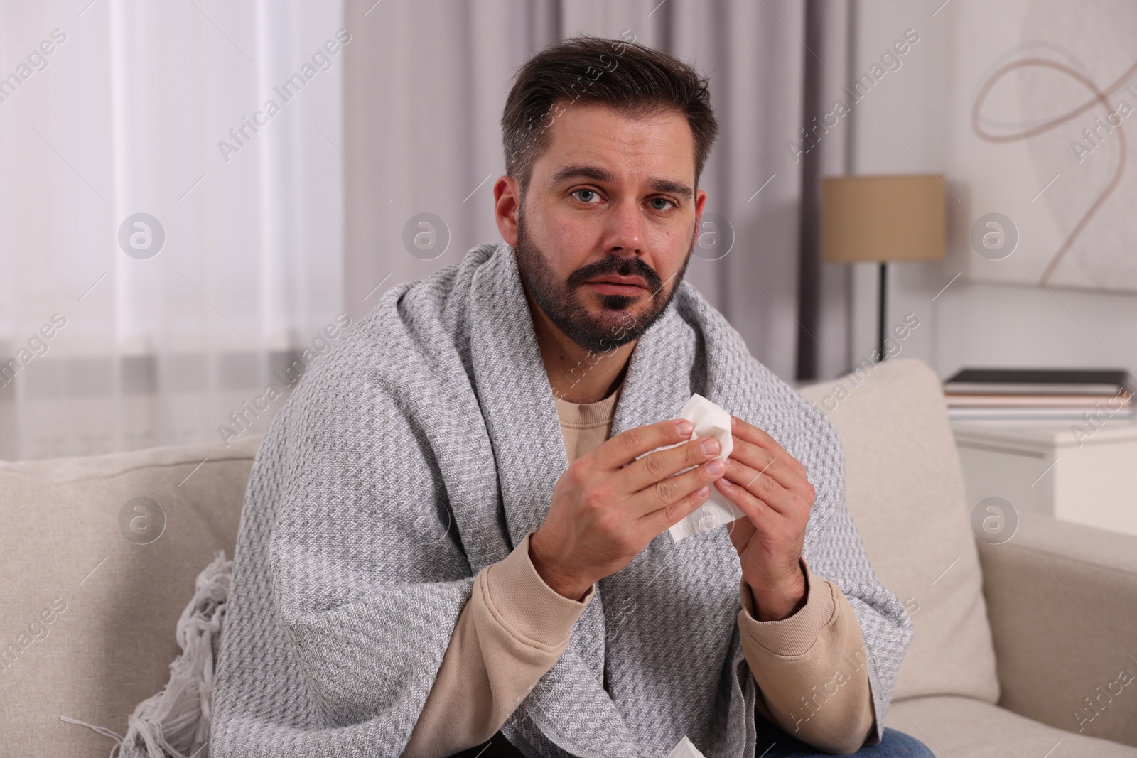 Photo of Sick man wrapped in blanket with tissue on sofa at home. Cold symptoms