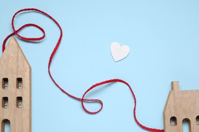 Photo of Red decorative cord and paper heart between two wooden house models on light blue background symbolizing connection in long-distance relationship, flat lay