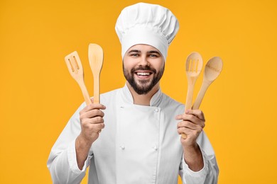 Photo of Happy young chef in uniform holding wooden utensils on orange background