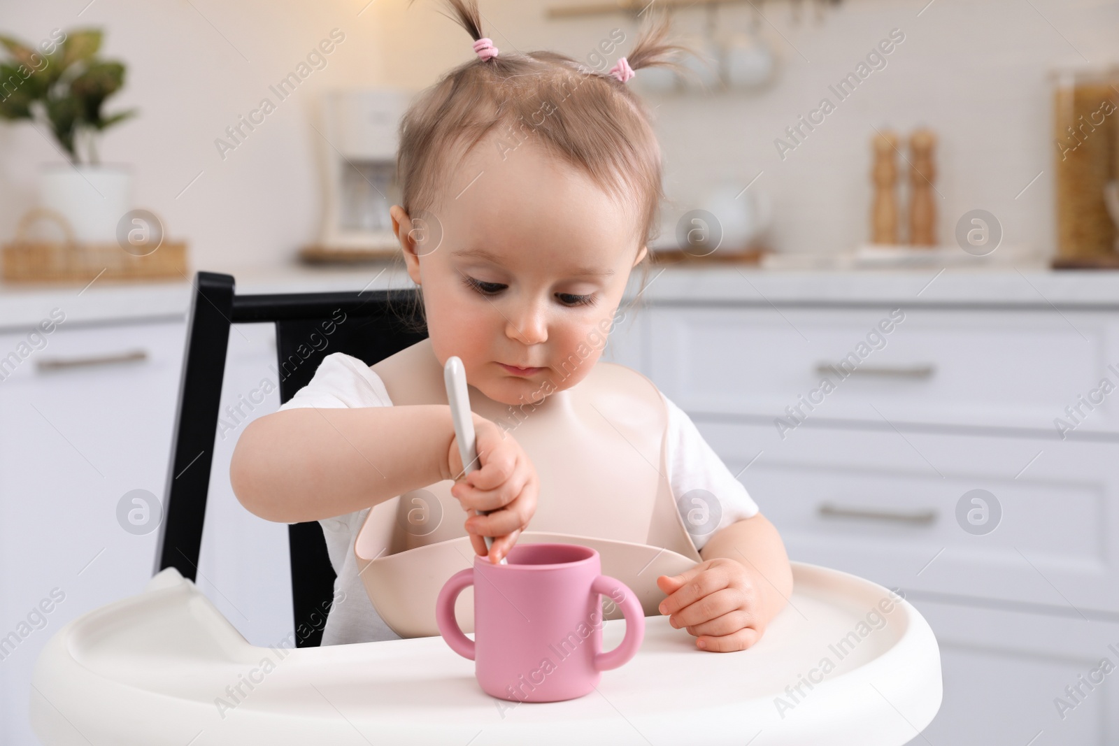 Photo of Cute little baby with spoon and cup in high chair indoors