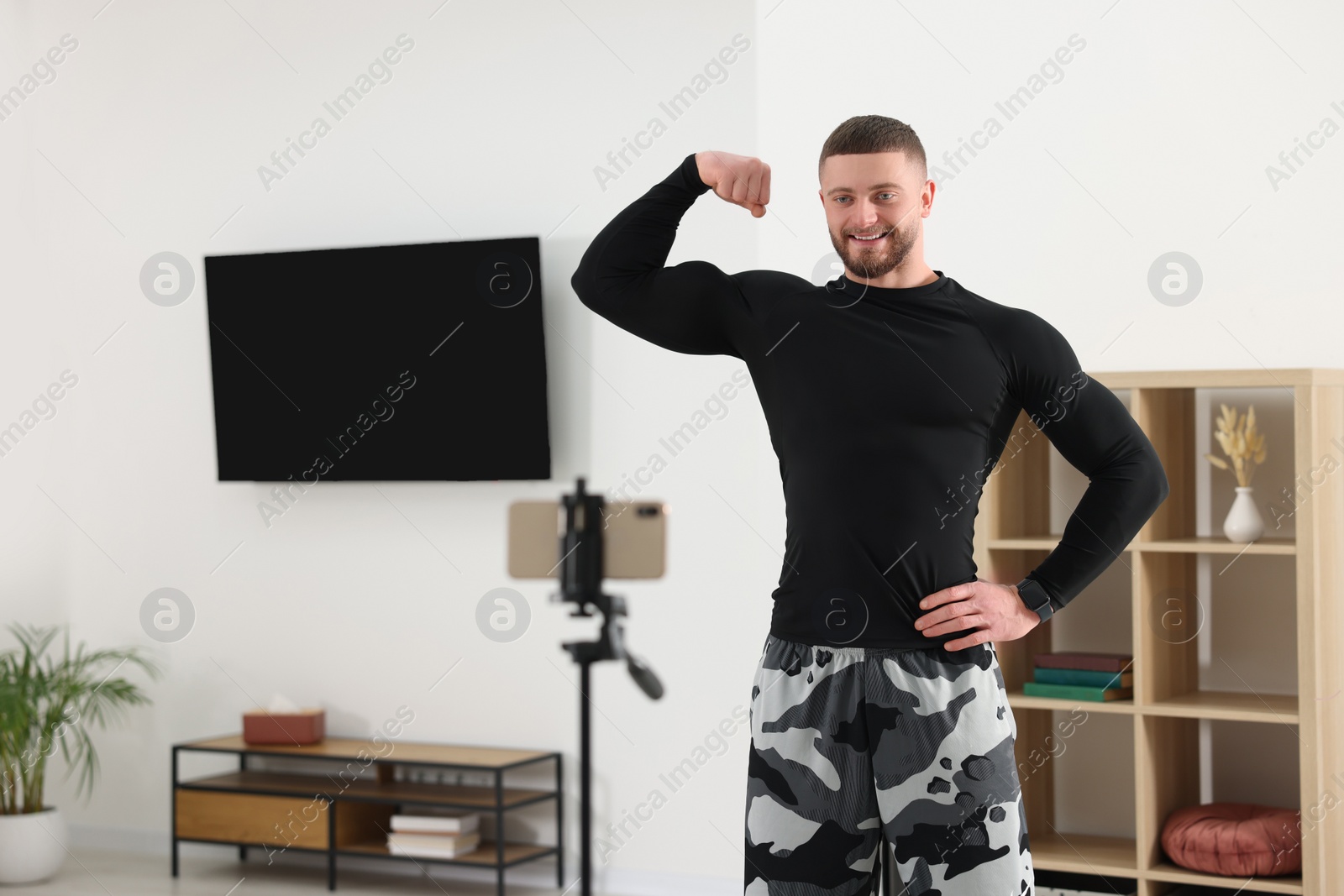 Photo of Trainer showing his muscles while streaming online fitness lesson with phone at home