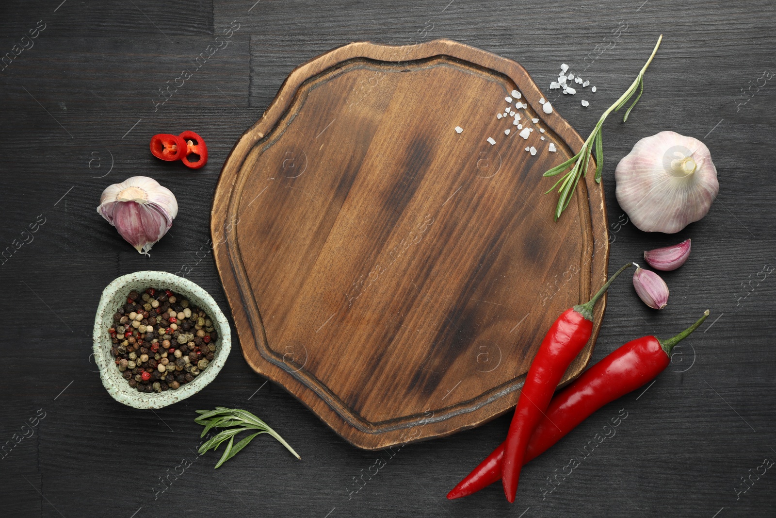 Photo of Cutting board, spices, garlic, rosemary and chili peppers on black wooden table, flat lay. Space for text