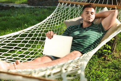 Young man with laptop resting in comfortable hammock at green garden