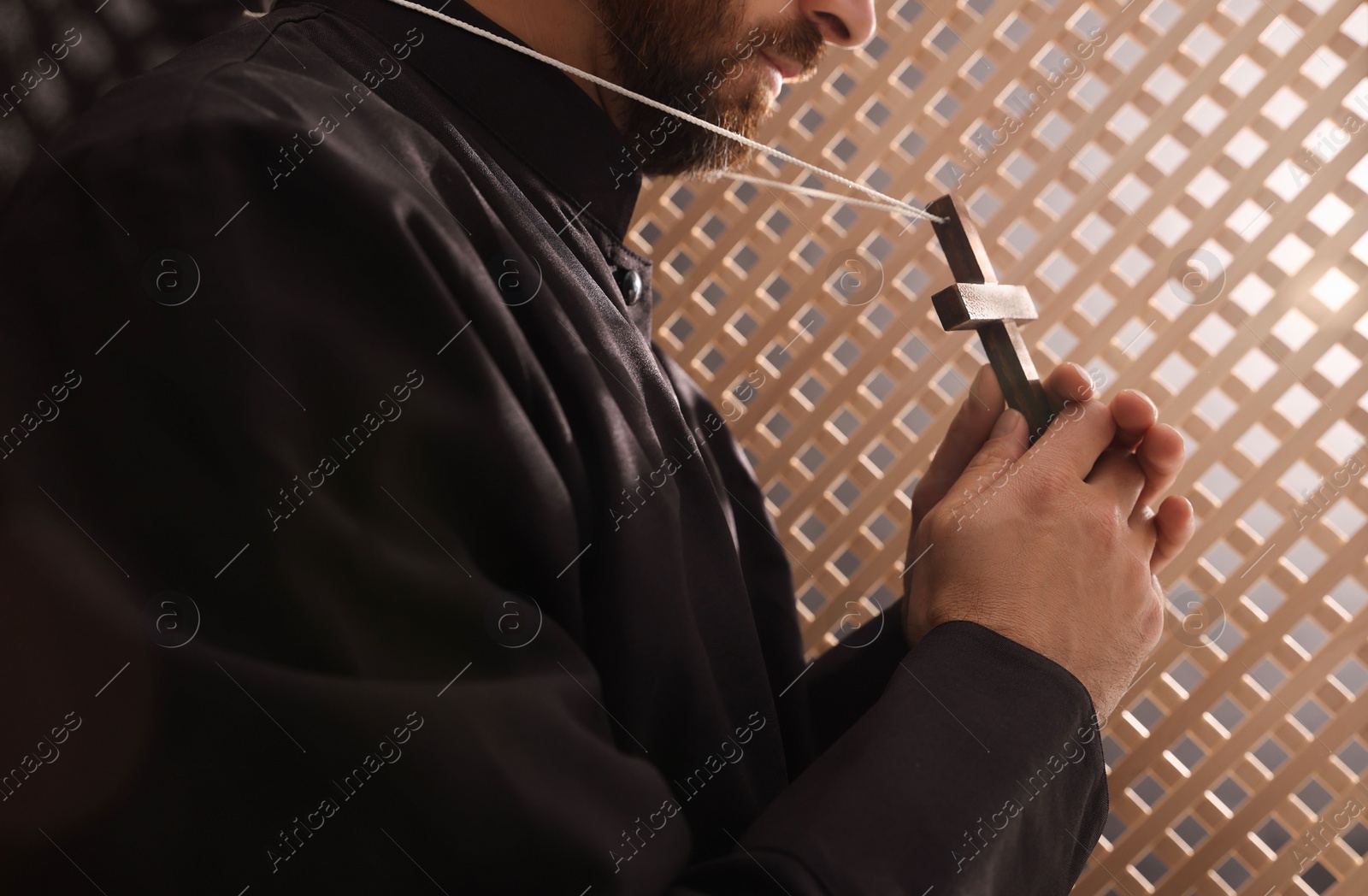 Photo of Catholic priest in cassock holding cross in confessional booth, closeup