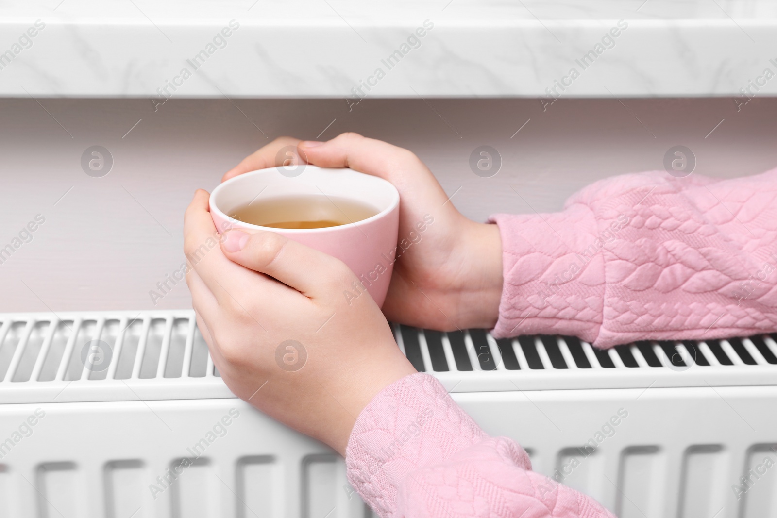 Photo of Girl with cup of tea warming hands on heating radiator indoors, closeup