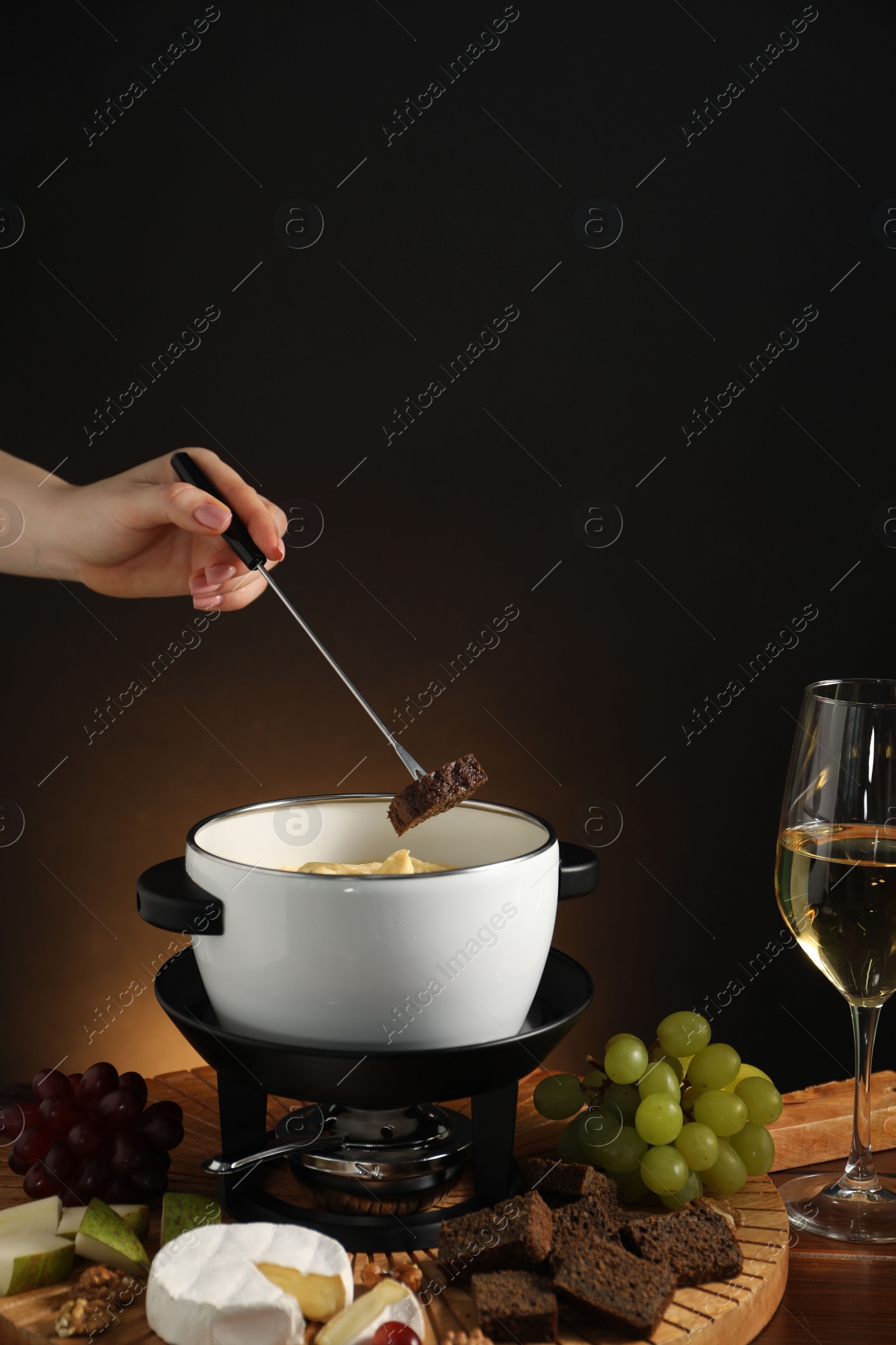 Photo of Woman dipping piece of bread into fondue pot with melted cheese at wooden table with wine and snacks, closeup