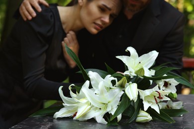 Photo of Sad couple mourning near granite tombstone with white lilies at cemetery outdoors, selective focus. Funeral ceremony