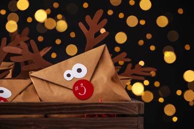 Photo of Gifts in envelopes with deer faces in wooden crate against blurred lights, closeup. Christmas advent calendar