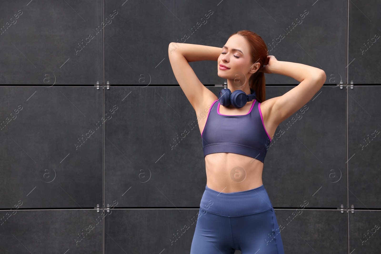 Photo of Beautiful woman in stylish gym clothes with headphones posing near dark grey wall on street, space for text