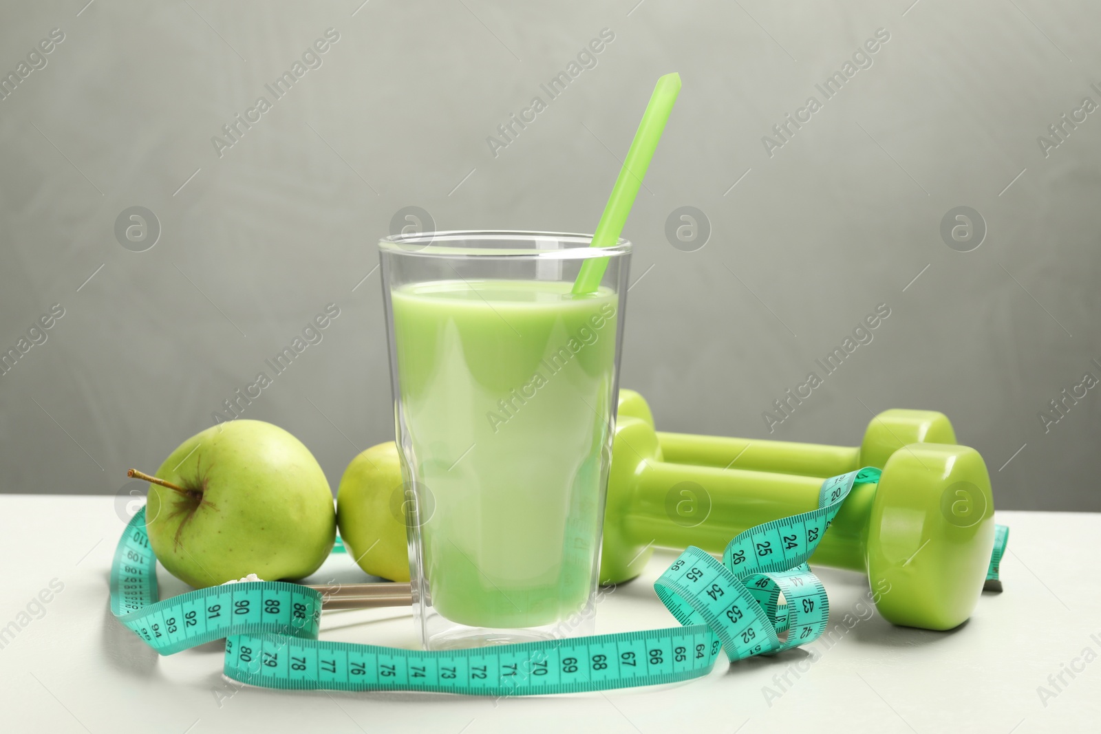 Photo of Tasty shake with apples, dumbbells and measuring tape on white table against gray background. Weight loss