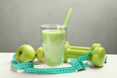 Tasty shake with apples, dumbbells and measuring tape on white table against gray background. Weight loss