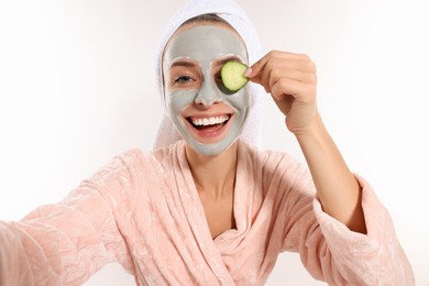 Photo of Woman with face mask and cucumber slice taking selfie on white background. Spa treatments