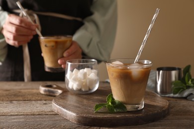 Photo of Glass of delicious iced coffee with milk on wooden table, selective focus. Woman stirring aromatic beverage with straw, closeup