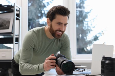 Professional photographer holding digital camera near table with laptop indoors