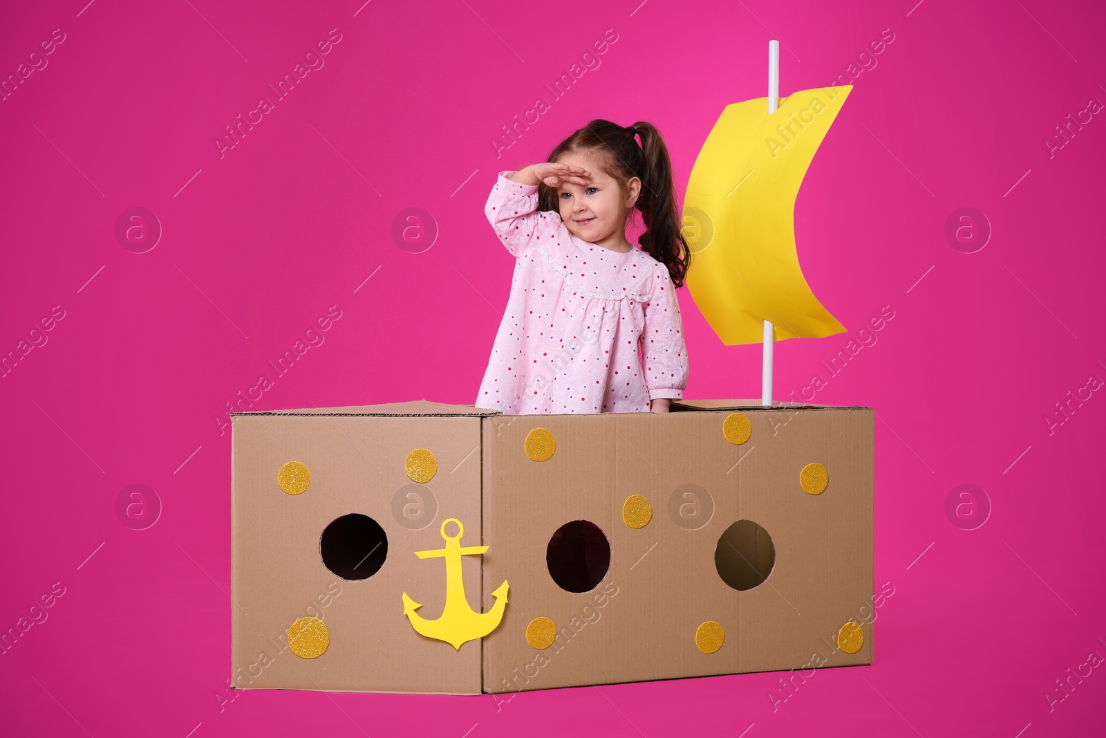 Photo of Little child playing with ship made of cardboard box on pink background