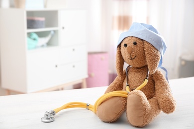 Photo of Toy bunny with medical cap and stethoscope on table indoors, space for text. Children's doctor