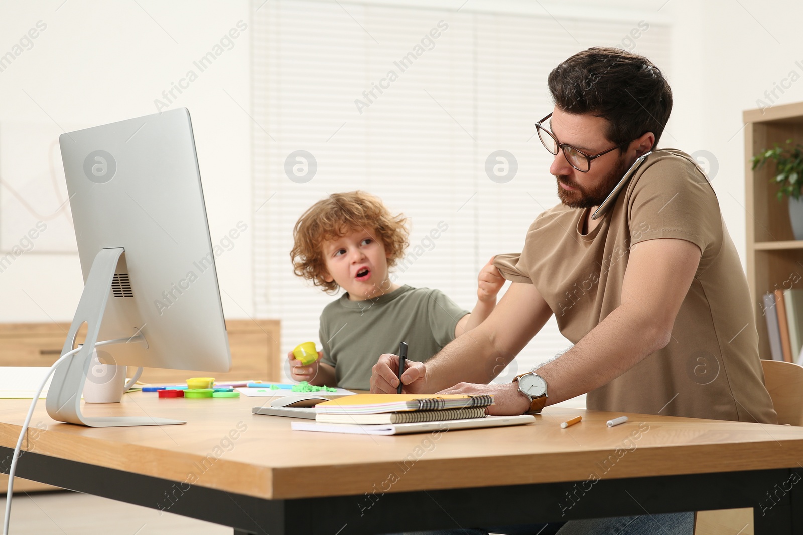 Photo of Little boy bothering his father at home. Man working remotely at desk