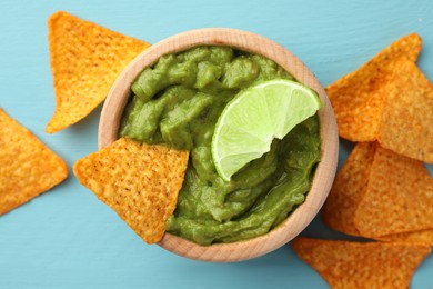 Bowl of delicious guacamole, lime and nachos chips on light blue wooden table, flat lay