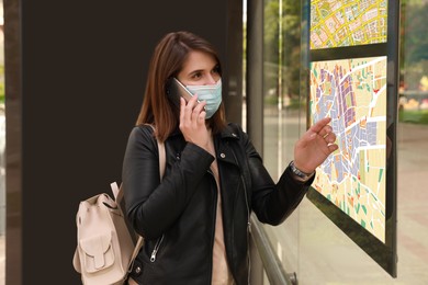 Image of Young woman in medical face mask talking on phone near public transport map at bus stop