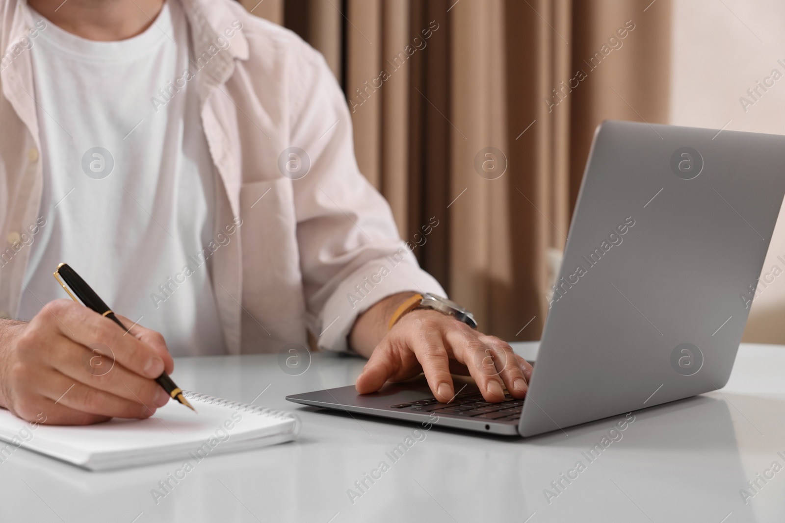 Photo of Man using laptop and writing something in notebook at table indoors, closeup