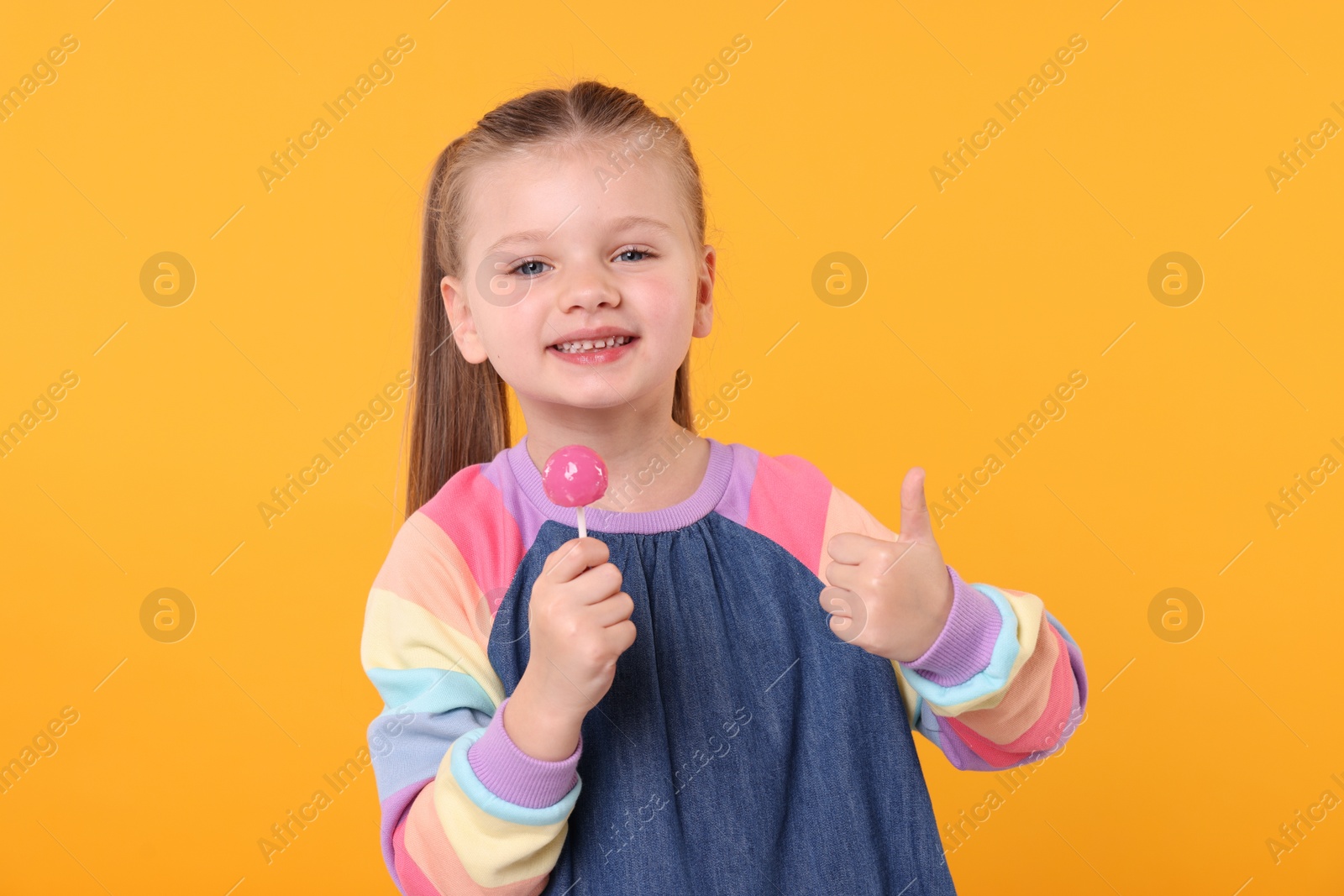 Photo of Happy little girl with lollipop showing thumbs up on orange background