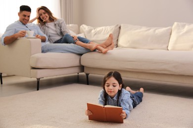 Photo of Little girl reading book while parents on sofa at home