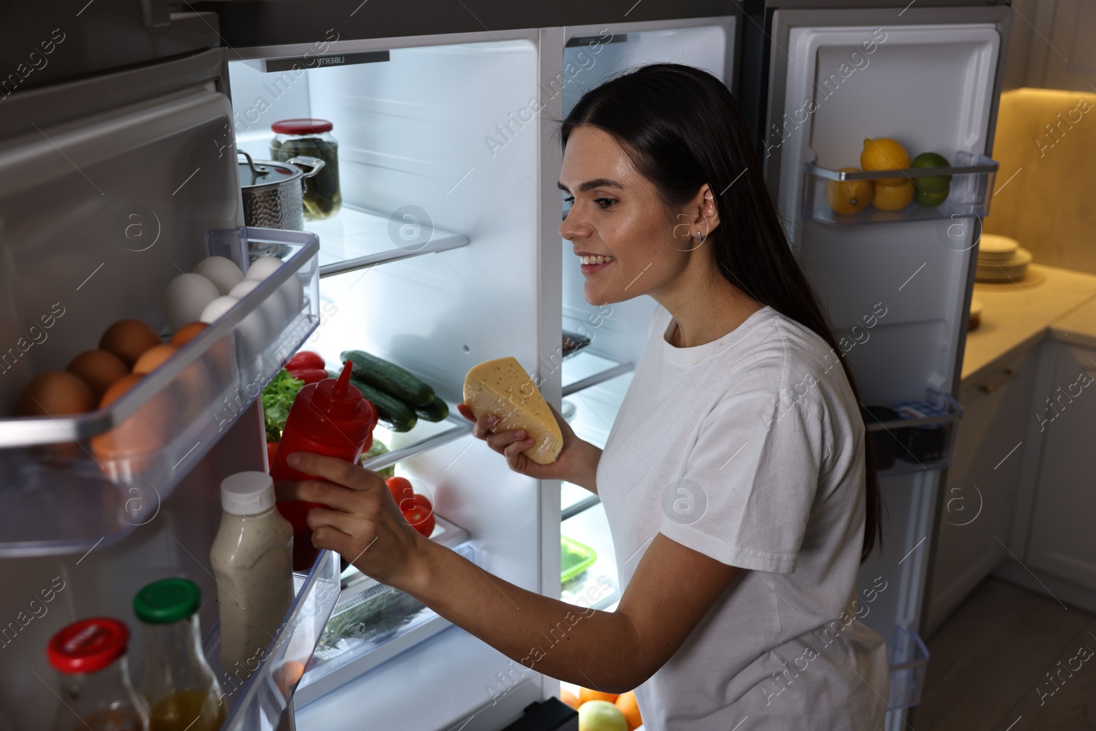 Photo of Young woman taking ketchup and cheese out of refrigerator in kitchen at night