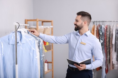 Photo of Dry-cleaning service. Happy worker with notebook choosing clothes from rack indoors