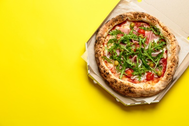 Photo of Tasty pizza with meat and arugula in cardboard box on yellow background, top view. Space for text