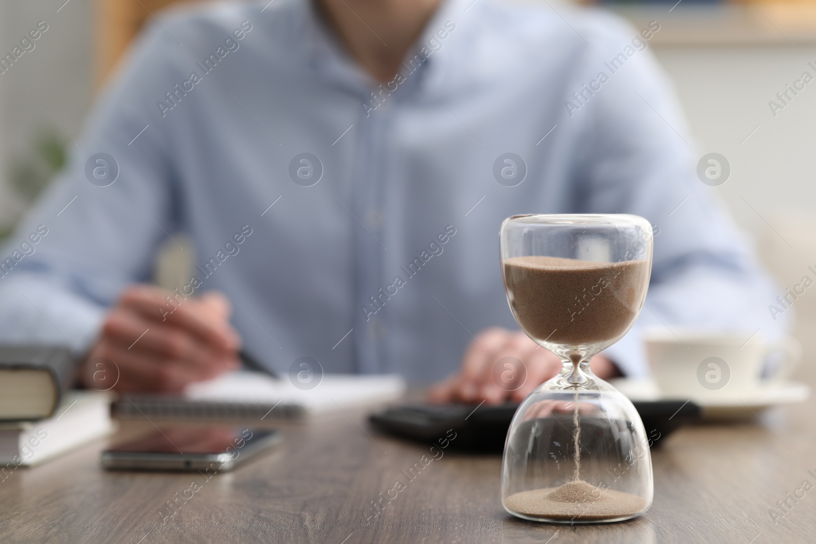 Photo of Hourglass with flowing sand on desk. Man taking notes while using calculator indoors, selective focus