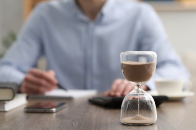 Hourglass with flowing sand on desk. Man taking notes while using calculator indoors, selective focus