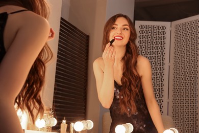 Photo of Beautiful young woman in elegant dress applying lipstick near mirror indoors
