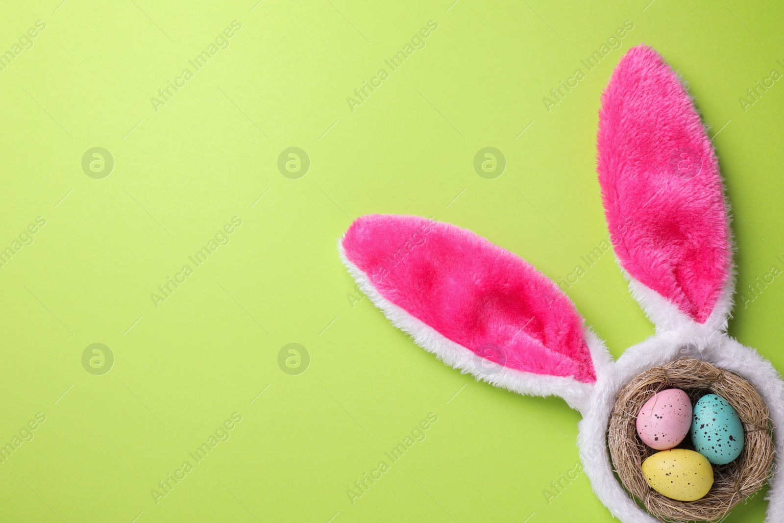 Photo of Headband with Easter bunny ears and dyed eggs in nest on green background, flat lay. Space for text