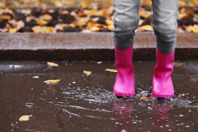 Photo of Little girl wearing rubber boots splashing in puddle on rainy day, focus of legs. Autumn walk