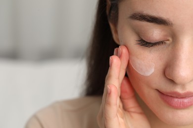 Young woman with dry skin applying cream onto her face against blurred background, closeup. Space for text