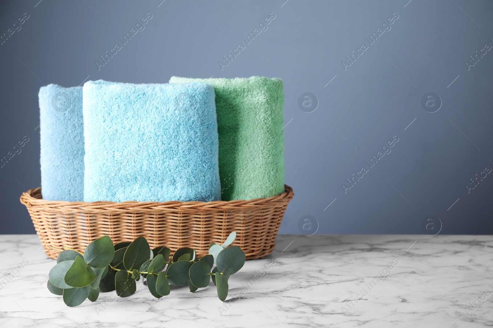 Photo of Wicker basket with folded bath towels and eucalyptus branch on white marble table. Space for text