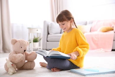 Photo of Cute little girl with teddy bear reading book on floor at home