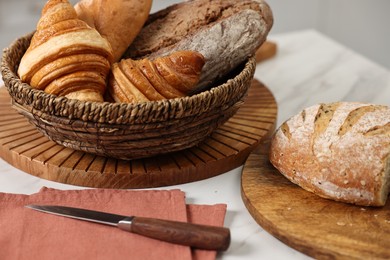 Photo of Wicker bread basket with freshly baked loaves and knife on white marble table in kitchen, closeup