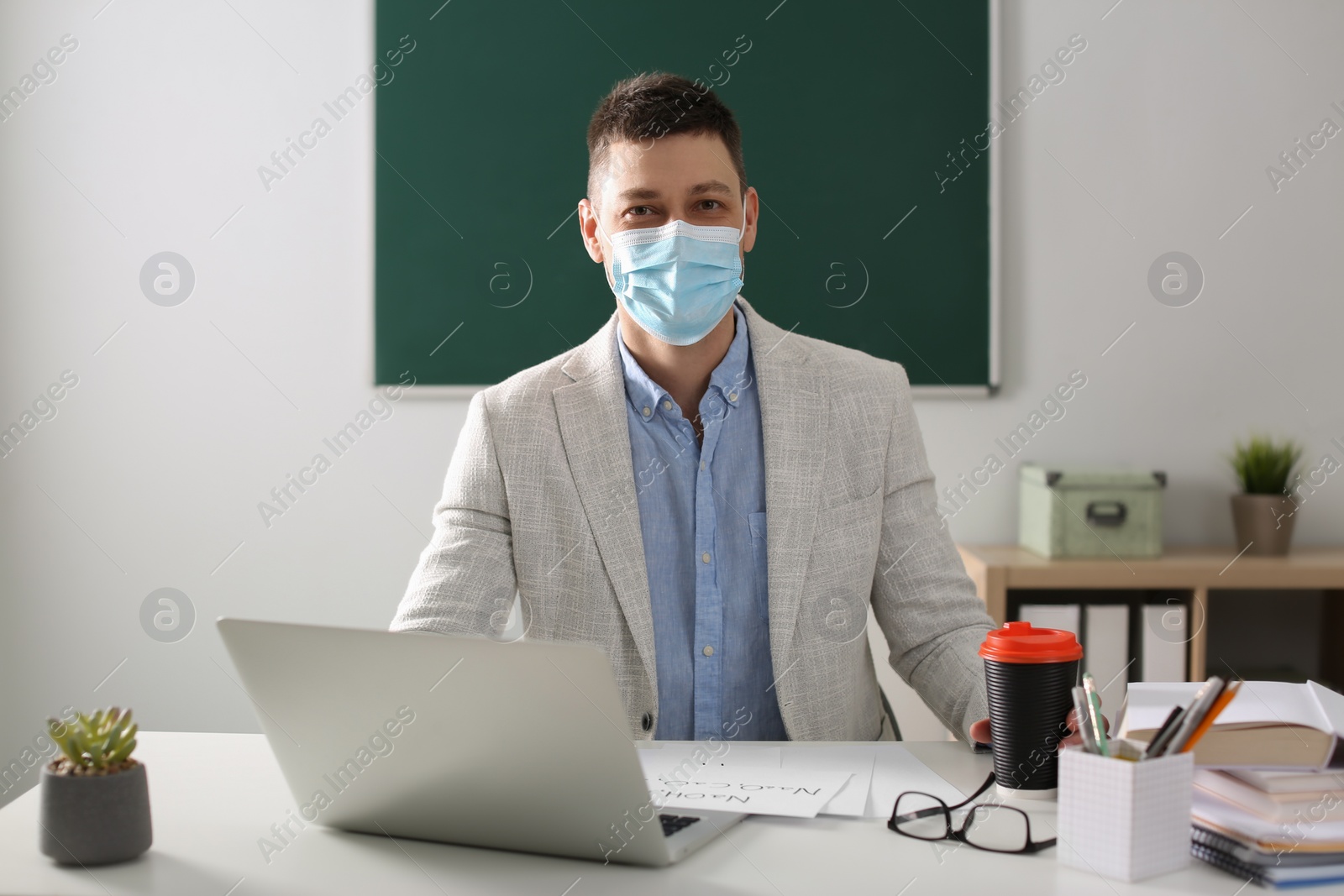 Photo of Teacher with protective mask and laptop sitting at desk in classroom. Reopening after Covid-19 quarantine