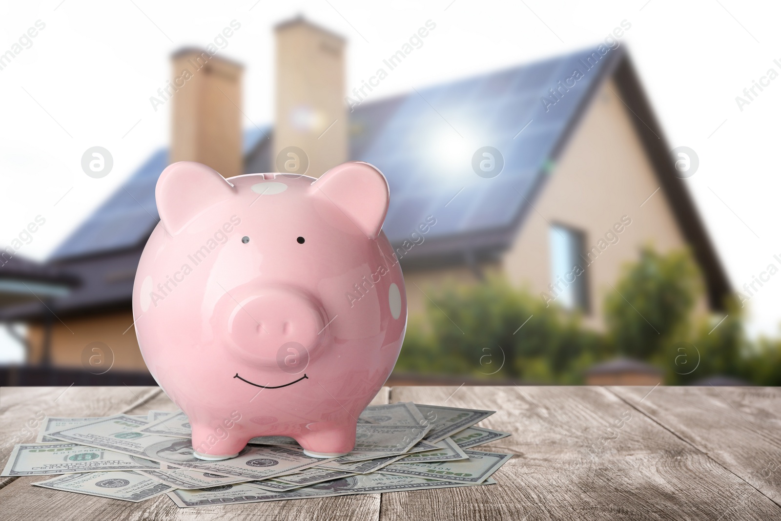 Image of Piggy bank and money on wooden surface and blurred view of beautiful house, space for text. Mortgage concept