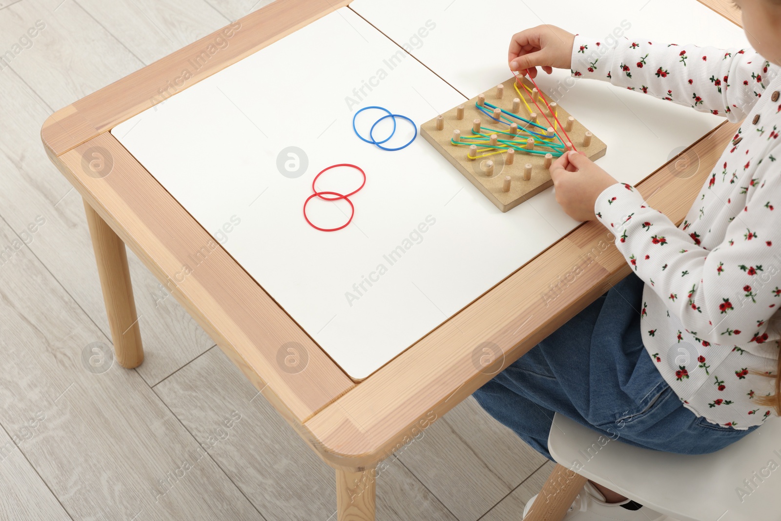 Photo of Motor skills development. Girl playing with geoboard and rubber bands at white table indoors, closeup. Space for text