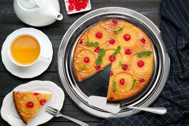 Photo of Delicious cut pineapple pie with cherry and tea served on black wooden table, flat lay