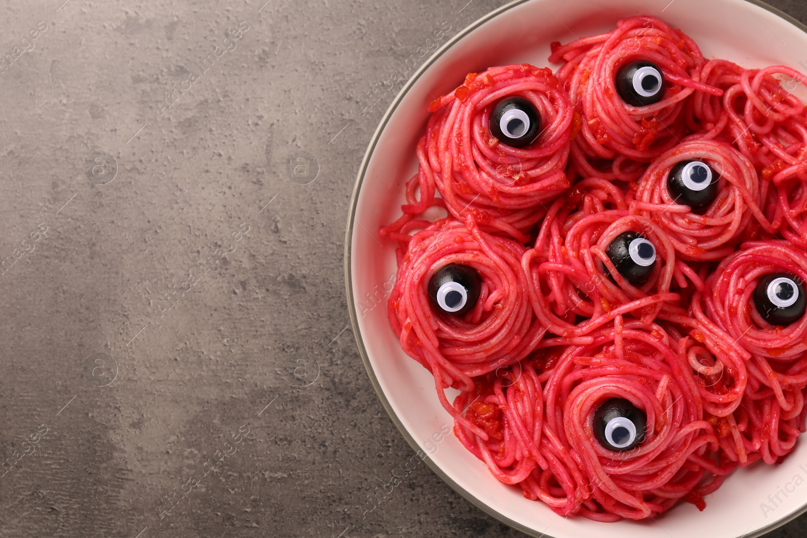 Photo of Red pasta with decorative eyes and olives in bowl on grey textured table, top view with space for text. Halloween food