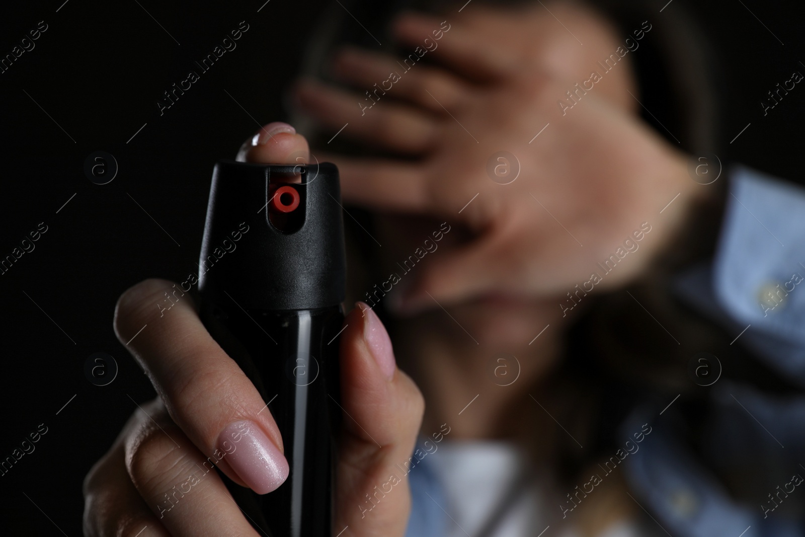 Photo of Young woman covering eyes with hand and using pepper spray on black background, focus on canister