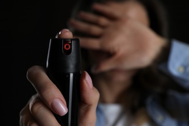 Photo of Young woman covering eyes with hand and using pepper spray on black background, focus on canister