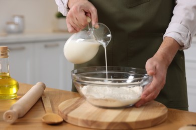 Photo of Making bread. Man pouring milk into bowl with flour at wooden table in kitchen, closeup
