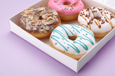 Photo of Box with different tasty glazed donuts on violet background, closeup